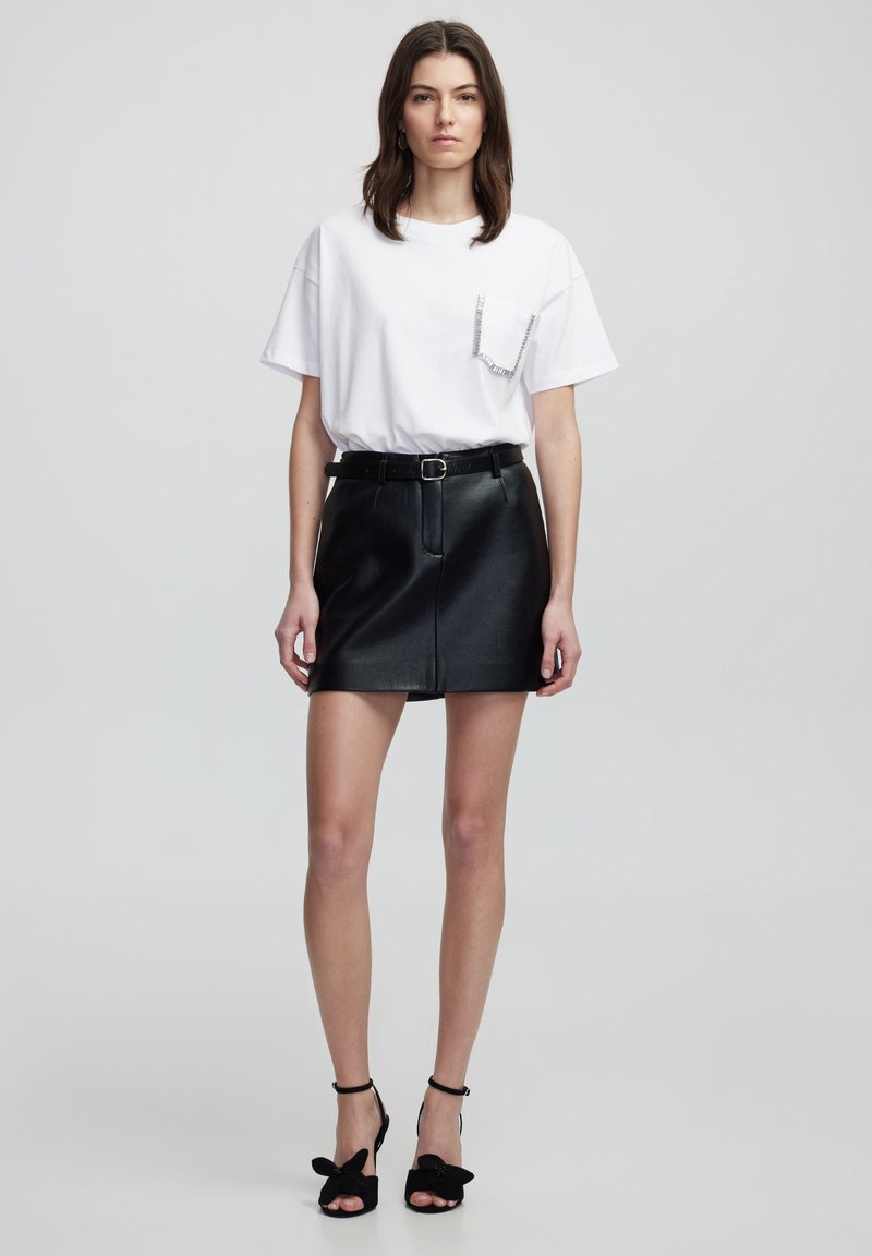 Weißes T-Shirt mit einem Strassdetail an der Tasche, kombiniert mit einem schwarzen Leder-Minirock und schwarzen High Heels mit Schleifenakzenten.
