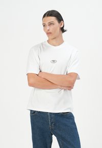 Un tânăr cu părul întunecat și pieptănat înapoi stă cu brațele încrucișate, purtând un tricou alb cu un mic logo negru și blugi albaștri.