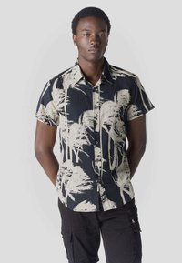 Jeune homme portant des boucles d'oreilles, une chemise noire à manches courtes avec un imprimé de palmiers beige et un short cargo noir, debout les mains derrière le dos.