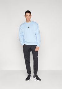 Lys blå sweatshirt med sort logo, parret med sorte sportbukser med hvid branding. Modellen har sorte atletiske sko på.