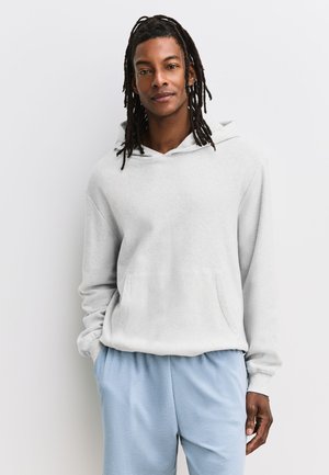 Homme avec des dreadlocks portant un sweat à capuche gris clair et un pantalon bleu clair, debout les mains dans les poches devant un mur uni.