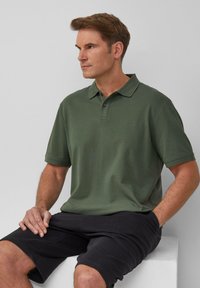 Donkergroen poloshirt met een geknoopte kraag, korte mouwen en geborduurd logo; gecombineerd met zwarte shorts, zittend tegen een lichte achtergrond.
