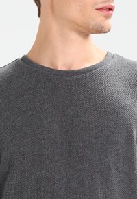 Suéter gris texturizado con cuello redondo, que presenta un sutil patrón tejido y puños acanalados. Primer plano que muestra el detalle de la tela.
