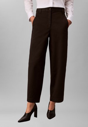 Mujer con pantalones negros de pierna ancha, zapatos negros de tacón alto y una camisa blanca de manga larga con botones, con las manos en los bolsillos.