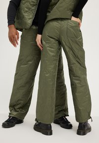 Pantalons matelassés verts olive avec une texture lisse, présentant une coupe décontractée, des poches latérales et des liens au niveau des chevilles.