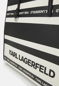 Sac cabas à rayures noires et blanches en toile, avec logo embossé. Comprend des accents en cuir et une poignée supérieure pour le transport.