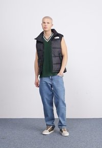 Gilet imbottito nero sopra un maglione verde a coste, jeans baggy blu chiaro e sneakers beige con accenti marroni. Stile semplice e casual.