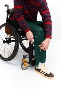 Személy ül a kerekesszékben, zöld nadrágon patentokat rögzít, csíkos pulóvert és fehér-barna tornacipőt visel.