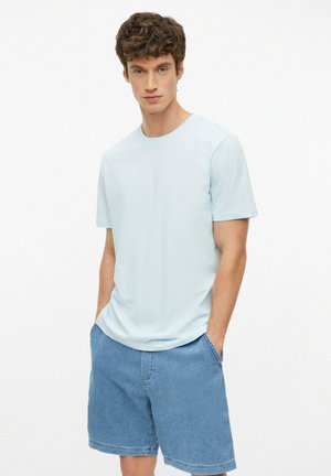 Młody mężczyzna z krótkimi kręconymi brązowymi włosami, ubrany w jasnoniebieski t-shirt i jeansowe szorty, stoi z rękami w kieszeniach na białym tle.