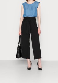 Donna con top senza maniche blu, pantaloni neri a gamba larga a vita alta con cintola, tacchi neri e che tiene una borsa nera.