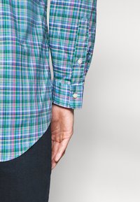 Arm und Hand einer Person neben dem Oberkörper, die ein blau-grün-lila kariertes Hemd mit Knöpfen und eine dunkle Hose trägt, vor einfarbigem Hintergrund.