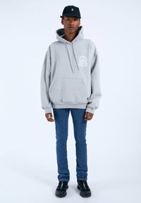 Felpa con cappuccio grigia, dotata di tasca frontale e dettaglio con logo, abbinata a jeans blu slim-fit e scarpe da ginnastica nere con accenti colorati.