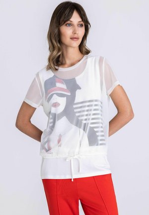 Femme aux cheveux mi-longs portant un haut graphique blanc transparent par-dessus une chemise blanche, associé à un pantalon rouge vif, debout devant un fond clair.