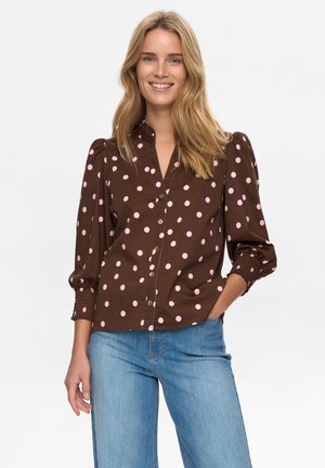 Braunes Hemd mit Knopfleiste und pinken Punkten, gerafften Ärmeln und lockerem Schnitt. Wird mit hellblauen Jeans getragen, verfügt über einen Stehkragen.