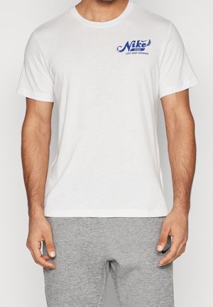 Mężczyzna w białej koszulce z okrągłym dekoltem z niebieskim napisem "Nike Seeds Just Keep Growing" na piersi, w połączeniu ze szarymi dresami.