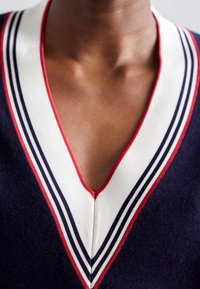 Gros plan d'une personne portant un pull bleu marine à col en V avec un liseré rayé blanc, rouge et noir autour de l'encolure.