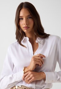 Witte button-up shirt met een klassieke kraag en lange mouwen, voorzien van parelknopen en een gladde textuur. Draagt een decoratieve gouden armband.