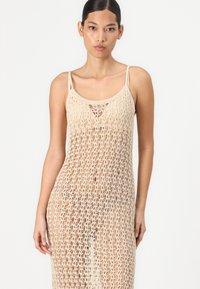 Beige crochet maxi dress with intricate open weave pattern, adjustable spaghetti straps, and a straight-cut design. Light and airy texture.
