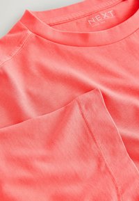 T-shirt en coton rose corail avec un col rond et des coutures visibles aux épaules et aux manches, étiqueté « NEXT Elbow Tex. »