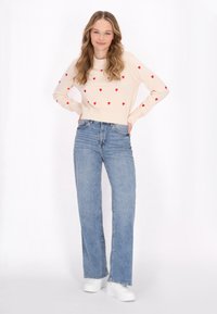 Maglione beige con motivi a cuore rossi, polsini e fondo a costine, abbinato a jeans dritti a vita alta blu chiaro e scarpe bianche.