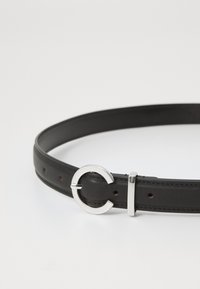 Ceinture en cuir noir avec une boucle en argent circulaire et un accent métallique droit. Surface texturée avec plusieurs trous de réglage.