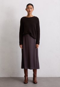 Bruine geribbelde gebreide trui, grijze satijnen midi-rok en bruine leren laarzen tot de knie, wat een gelaagde, monochrome outfit creëert.