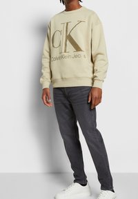 Sudadera color crema con gran logo beige "CK", escote redondo, puños acanalados, combinada con jeans gris oscuro y zapatillas blancas.