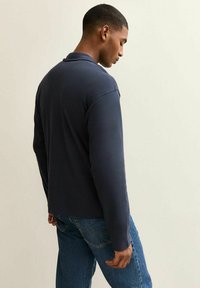 Polo a maniche lunghe blu navy con colletto classico, realizzato in tessuto morbido; abbinato a jeans in denim azzurro, che mostrano una vestibilità casual.