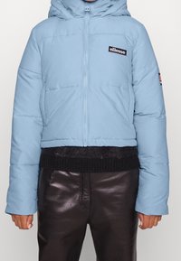 Jasnoniebieska kurtka puchowa z zamek błyskawicznym i kapturem, z czarną łatą logo na piersi i prążkowanym czarnym wykończeniem, noszona na błyszczących czarnych spodniach.
