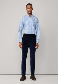 Man in een blauw geruit overhemd met lange mouwen, marineblauwe corduroypantalon en bruine dresschoenen, staand tegen een effen lichtgrijze achtergrond.