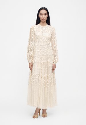 Femme aux longs cheveux foncés portant une robe en dentelle crème à manches longues avec une broderie florale et des chaussures beiges ouvertes à talons hauts.