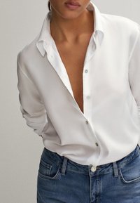 Valkoinen nappikauluspaita, jossa on syvä pääntie, valmistettu sileästä kankaasta. Yhdistetty tummansinisiin farkkuihin, joissa on näkyviä metallinappitietoja.