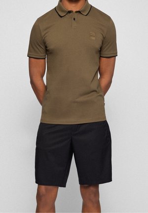 Oliwkowa koszulka polo z czarnymi akcentami, krótkie rękawy i kwadratowa naszywka z logo. Do tego czarne szorty o gładkiej fakturze.
