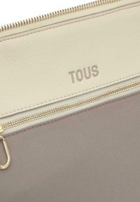 Funda de cuero beige con un zipper dorado, que presenta una base de tela texturizada y el logo "TOUS" en relieve en la parte frontal.