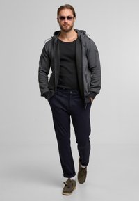 Felpa grigia con zip e texture a fantasia, top nero, pantaloni blu e scarpe marroni con lacci. Gli occhiali da sole completano l'outfit.