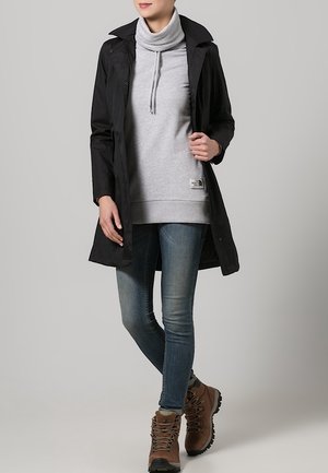 Kobieta ubrana w czarny płaszcz, szarą bluzę z golfem, obcisłe dżinsy i brązowe buty trekkingowe stoi na jednolitym tle.