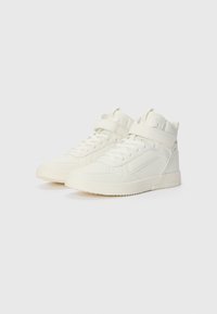 Baskets blanches montantes en cuir lisse avec un bout perforé, une cheville rembourrée et une sangle velcro au niveau du métatarse.
