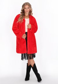 Palton faux fur roșu, cu mâneci lungi, deschis în față și o croială lejeră, asortat cu o fustă neagră cu franjuri și cizme negre din suede.