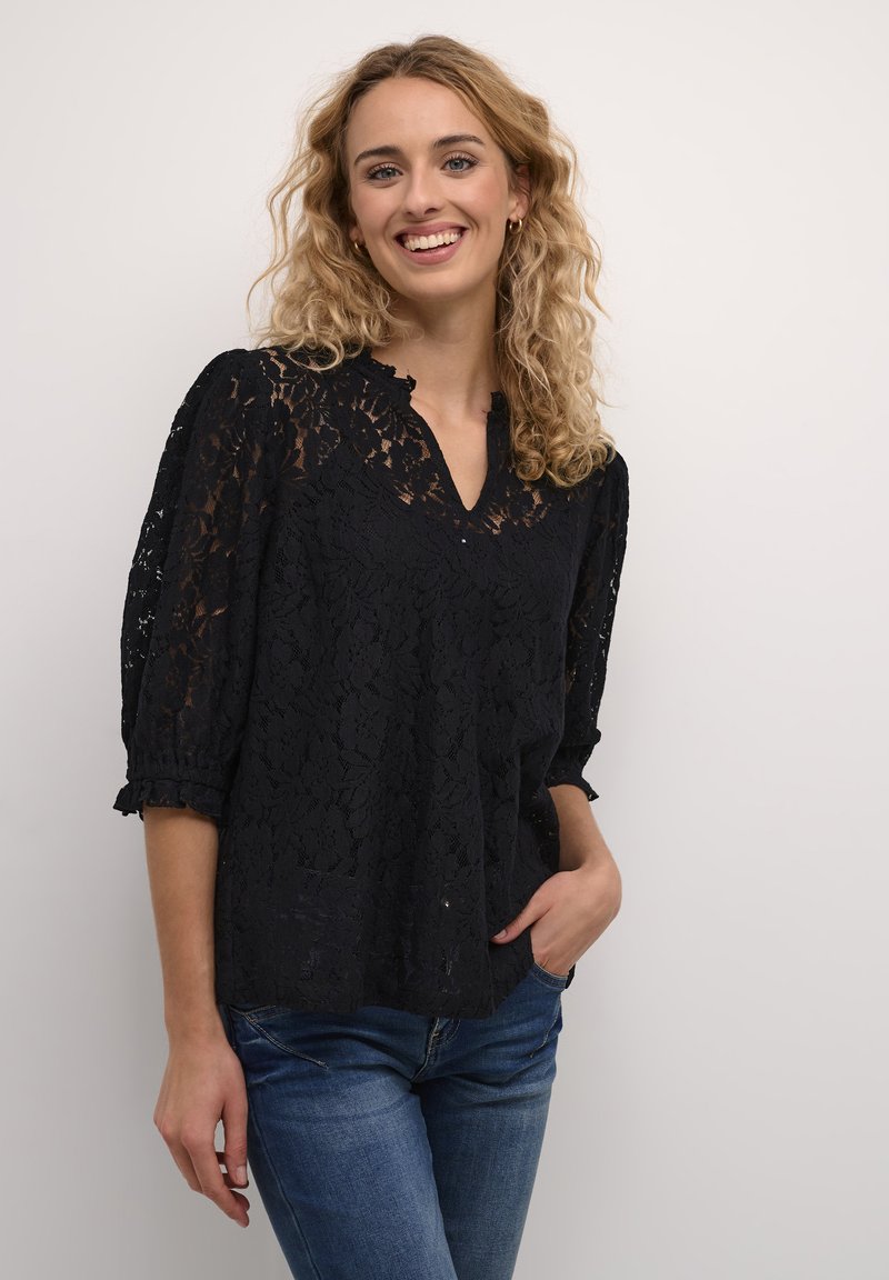 Black lace blouse with a V-neck, puffed sleeves, and ruffled cuffs. The fabric features a floral pattern and paired with blue jeans.