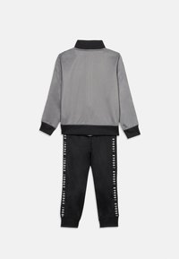 Γκρι αθλητικό μπουφάν με μαύρο γιακά και μανσέτες, συνδυασμένο με μαύρο παντελόνι που έχει κατακόρυφο λευκό μοτίβο λογότυπου στις πλευρές. Ομαλή υφή.