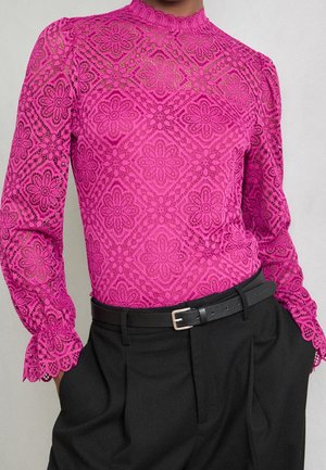 Haut en dentelle à manches longues rose vif avec des motifs floraux, associé à un pantalon noir taille haute et une ceinture noire.