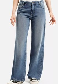 Kobieta w niebieskich, wysokich, szerokich dżinsach i czarno-białych trampkach, stoi z rękami swobodnie opuszczonymi wzdłuż ciała.