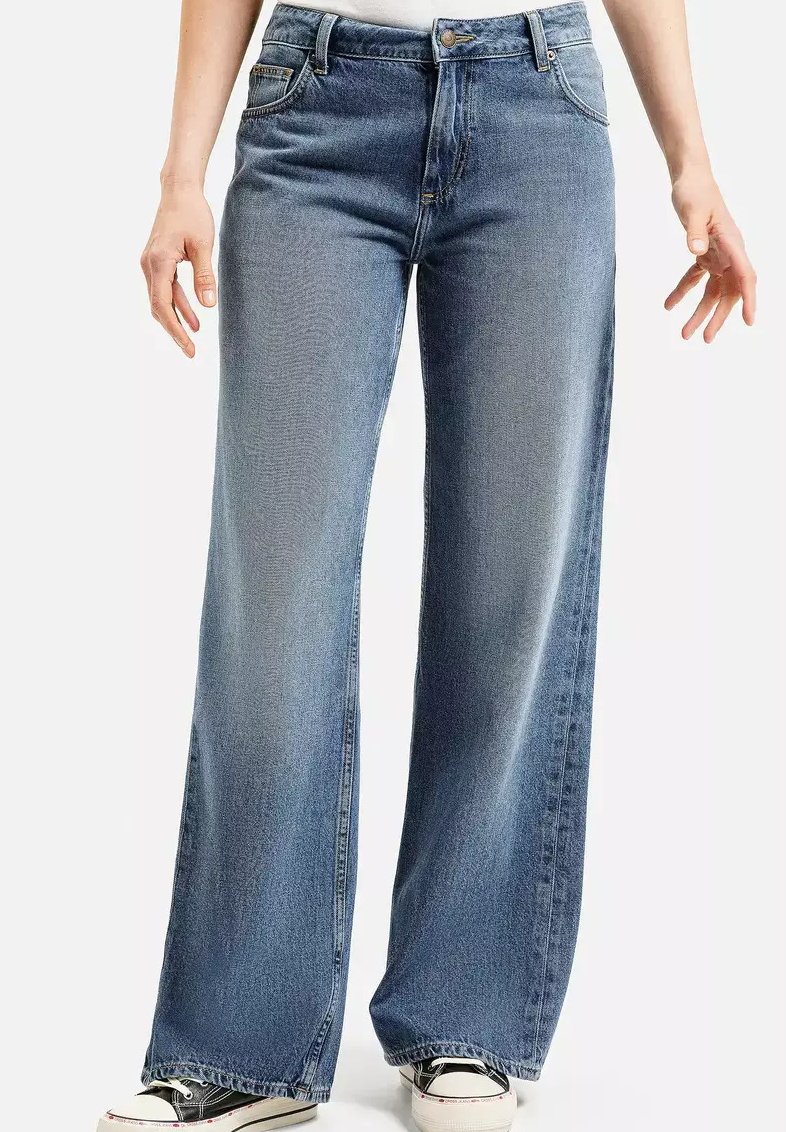 Kobieta w niebieskich, wysokich, szerokich dżinsach i czarno-białych trampkach, stoi z rękami swobodnie opuszczonymi wzdłuż ciała.