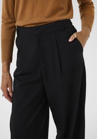 Black tailored trousers with pleats and side pockets, paired with a brown knit sweater. Smooth fabric with a subtle sheen.