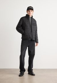 Zwarte pufferjack met horizontale gestikte desing, hoge kraag en logo patch, gecombineerd met donkere jeans en zwarte schoenen.