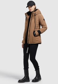 Bruine gewatteerde jas met een capuchon, zwarte accenten en ritsen. Gdragend met een zwart topje en leggings, en zwarte enkellaarsjes.