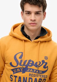 Giovane uomo che indossa un felpa gialla con il testo blu "Super Dry Japan Registered Standard", sovrapposta a una camicia a quadri, con espressione neutra.