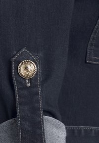 Gros plan sur un tissu en denim foncé avec un revers plié et un bouton décoratif doré fixé à une lanière cousue.
