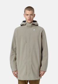 Cappotto impermeabile beige chiaro con colletto alto, cappuccio e logo argentato. Presenta un design slanciato e maniche lunghe. Texture liscia.