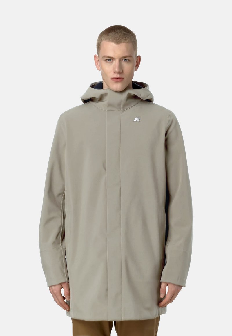 Cappotto impermeabile beige chiaro con colletto alto, cappuccio e logo argentato. Presenta un design slanciato e maniche lunghe. Texture liscia.